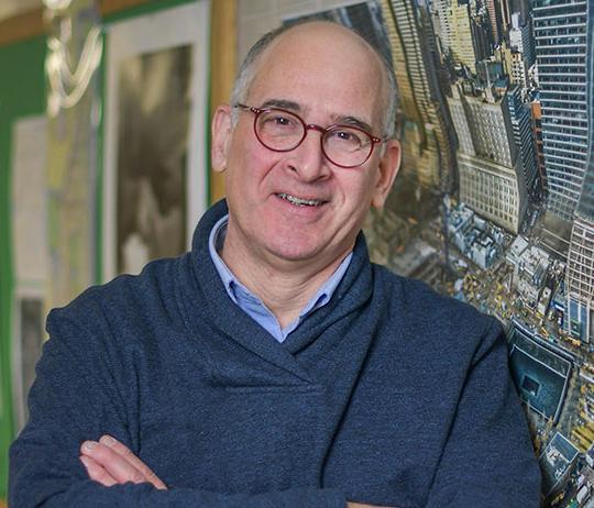 headshot of man with glasses, standing by poster of New York City